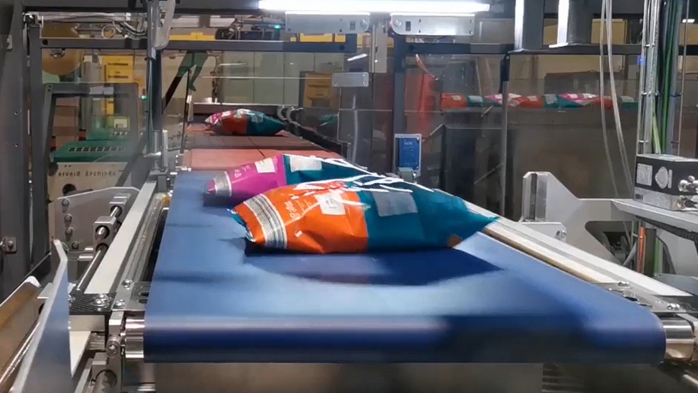 Colourful snack bags moving along a blue conveyor at an automated secondary packaging machine.